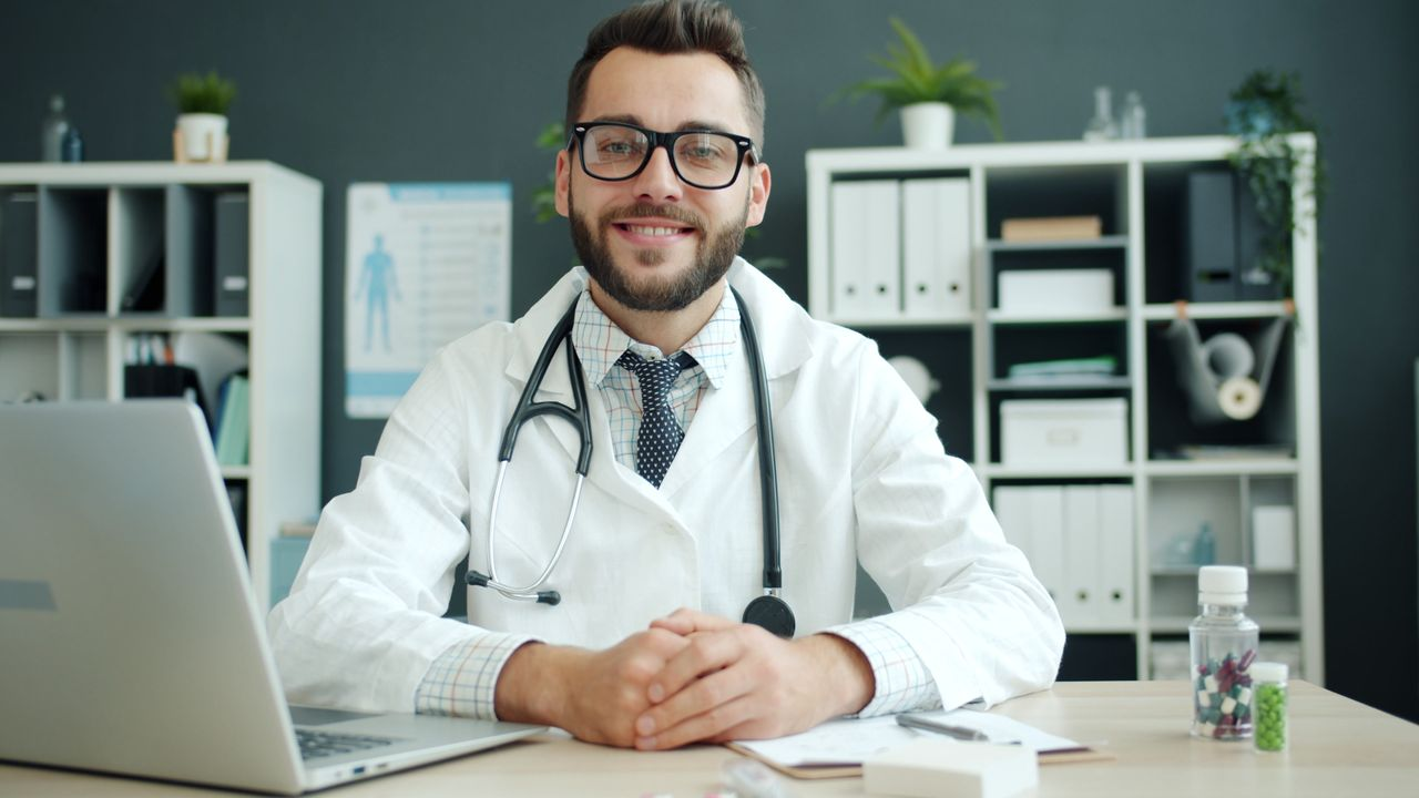 Friendly male doctor with stethoscope sitting at office desk in clinic.